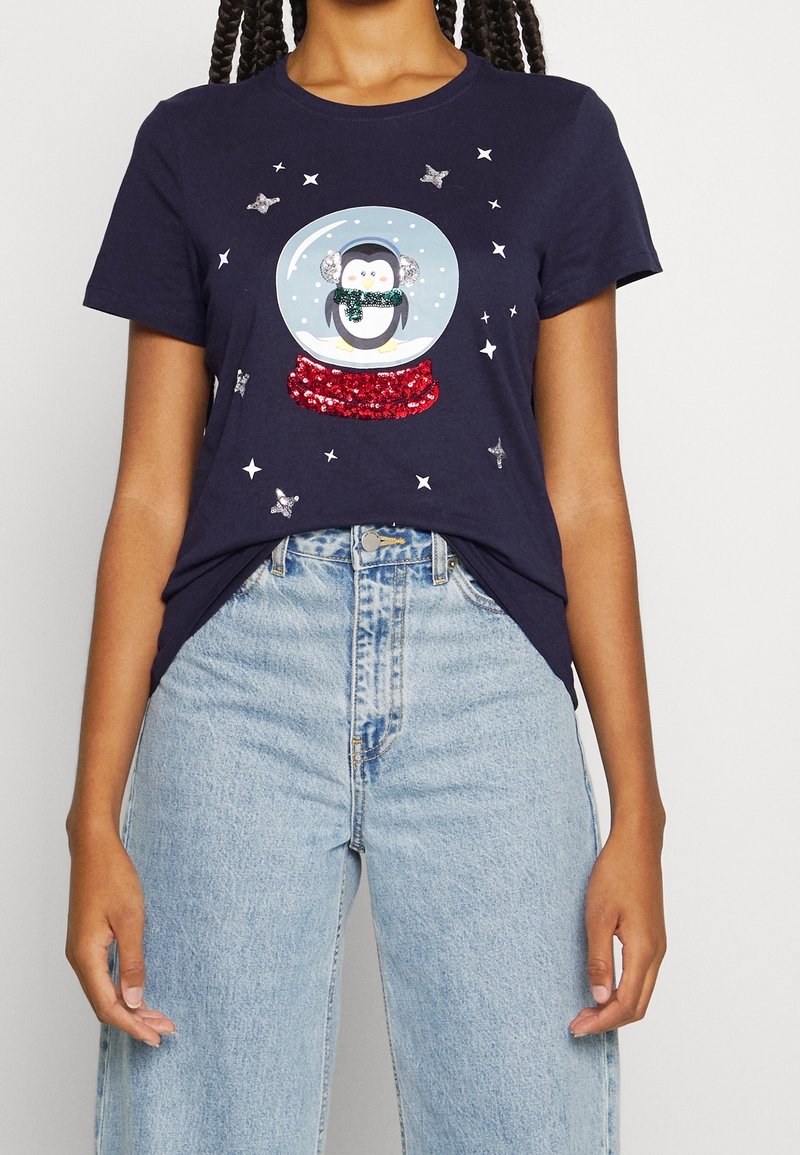Femme portant un t-shirt bleu marine avec un pingouin scintillant à l'intérieur d'un globe de neige, associé à un jean clair taille haute.