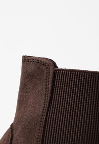 Botín de ante marrón con un panel textil acanalado, costuras contrastadas y un borde superior redondeado.