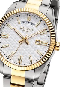 Montre en métal argenté et doré avec un cadran blanc texturé, des index dorés et un affichage de la date. Elle possède une lunette rainurée et un bracelet métallique.