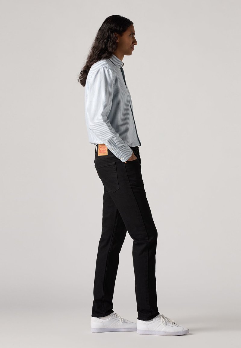 Chemise à rayures bleu clair à manches longues, associée à un jean noir et des baskets blanches. Détail de tag remarquable sur la poche arrière du jean.