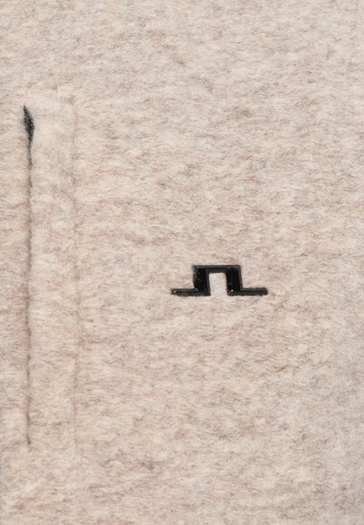 Beiges Wollgewebe mit einer weichen Textur und einem schwarzen Logo-Detail. Das Design umfasst eine gerade Tasche auf der linken Seite.