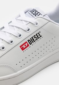 Vita syntetiska sneakers med perforerat mönster, med "DIESEL"-logga i svart och röd, och platta vita snören. Texturerad gummisul.