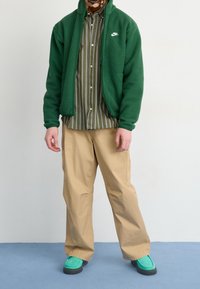 Vihreä fleecetakki raidallisen oliivinvihreän paidan päällä, beiget cargo-housut ja sinivihreät kengät harmailla pohjilla. Tekstuuriset materiaalit ja rento istuvuus.