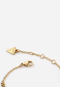 Bracciale in metallo dorato con catena, caratterizzato da un pendente triangolare con la scritta "Guess" e una chiusura a gancio. Maglie rotonde con una superficie liscia.
