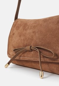 Brązowa torebka ze skóry zamszowej o gładkiej fakturze, z wiązanym zapięciem, cienkim skórzanym paskiem i złotymi ozdobami na końcach.