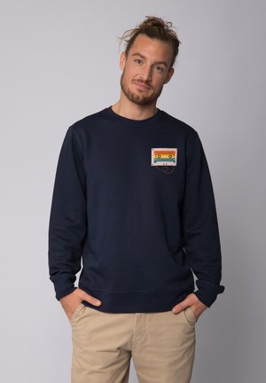 Navy sweatshirt with long sleeves and round neckline. Features a colorful cassette graphic on the chest pocket area. Soft fabric texture.