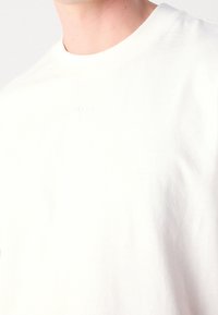 Witte katoenen t-shirt met een ronde hals. Heeft een subtiele reliëflogo op de borst. De textuur lijkt zacht en glad te zijn.