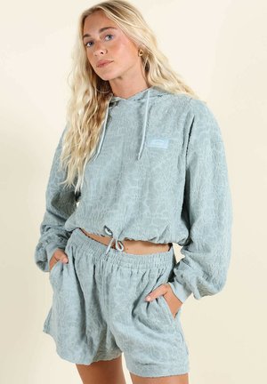 Jeune femme aux longs cheveux blonds portant un sweat à capuche texturé bleu clair et un short assorti, les mains dans les poches, debout devant un fond uni.