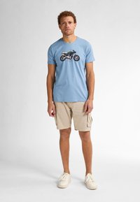 Lichtblauw katoenen t-shirt met een grafisch motief van een motorfiets, gecombineerd met beige cargoshorts en witte sneakers. Effen achtergrond.