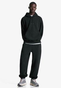 Haut à capuche noir avec poche kangourou et cordons, associé à un pantalon de survêtement noir assorti. L'ensemble est complété par des baskets grises.