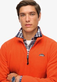 Suéter de forro polar naranja brillante con cuello con cremallera, que presenta una camisa de cuadros azul y blanca debajo. Logo "Superdry" en negro y acentos de herrajes.