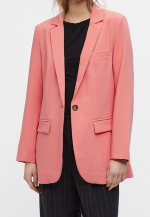 Personne portant un blazer rose corail par-dessus un haut noir et un pantalon sombre à fines rayures sur un fond clair uni.