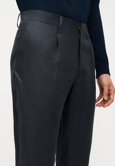 Άνδρας που φοράει σκούρο, ραμμένο παντελόνι και σκούρο πουλόβερ με μακριά μανίκια, στέκεται με το δεξί χέρι χαλαρό στο πλάι.