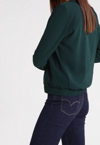 Mørkegrøn bomberjakke med ribbenede manchetter og kant, lavet af glat stof; parret med mørke jeans med design af baglomme.