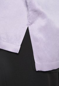 Camicia lilla con orlo e spacco laterale indossata sopra pantaloni neri, che mostrano la texture del tessuto e i dettagli delle cuciture.