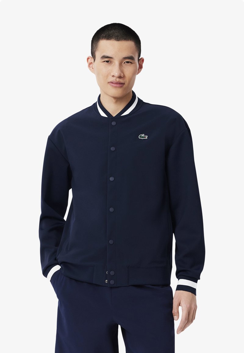 Chaqueta varsity azul marino con un diseño liso, cuello y puños acanalados que presentan rayas blancas, y un pequeño logo verde en el pecho.