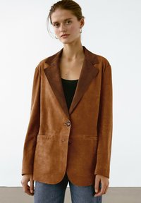 Brun läderblazer med slagkrage, enkel knappstängning, två framfickor och en slät textur, bärs över en svart topp.