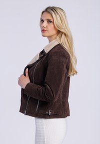 Model met lang blond haar, gekleed in een bruin suède jack met een schapenvachtkraag, poseert zijwaarts tegen een neutrale achtergrond.