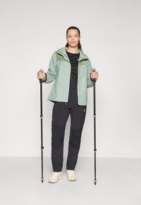 Veste imperméable vert clair avec capuche, chemise noire, pantalons de randonnée noirs et chaussures beiges, tenant des bâtons de trekking.