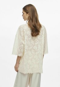 Top de encaje con un ajuste holgado, color crema, dobladillo festoneado y patrón floral; cuenta con mangas anchas y una textura ligera.