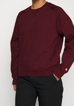 Homme portant un sweat-shirt bordeaux à manches longues avec poignets côtelés et pantalon noir, main dans la poche.