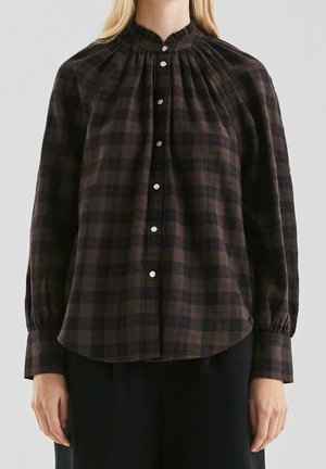 Femme portant un chemisier à carreaux marron et noir à manches longues avec des boutons argentés et un col froncé, associé à un pantalon noir.