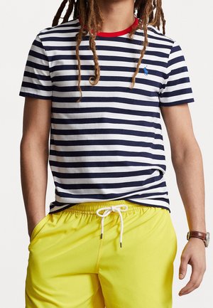 Person med dreadlocks iført en navy og hvidstribet t-shirt med rød krave og gule snørede shorts med hænderne i lommerne.