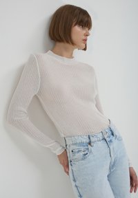Top lungo a maniche lunghe di un rosa chiaro a coste, con un collo alto, abbinato a jeans azzurri a vita alta, su uno sfondo bianco uniforme.