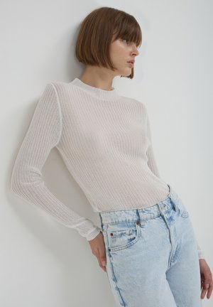 Top rose clair à manches longues côtelé avec un col montant, associé à un jean taille haute bleu clair, sur un fond blanc uni.