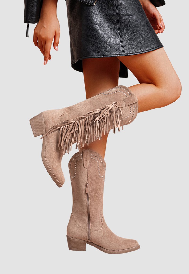 Femme portant des bottes de cow-boy en suédine beige avec des franges et des clous, associées à une jupe et une veste en cuir noires.