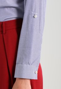 Blue and white striped shirt sleeve with a pearl button detail, paired with red trousers. The fabric is smooth with a tailored fit.