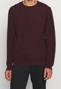 Vínový svetr s kulatým výstřihem v texturovaném pletení, s dlouhými rukávy a žebrovanými manžetami a lemem, nošený přes černé kalhoty.