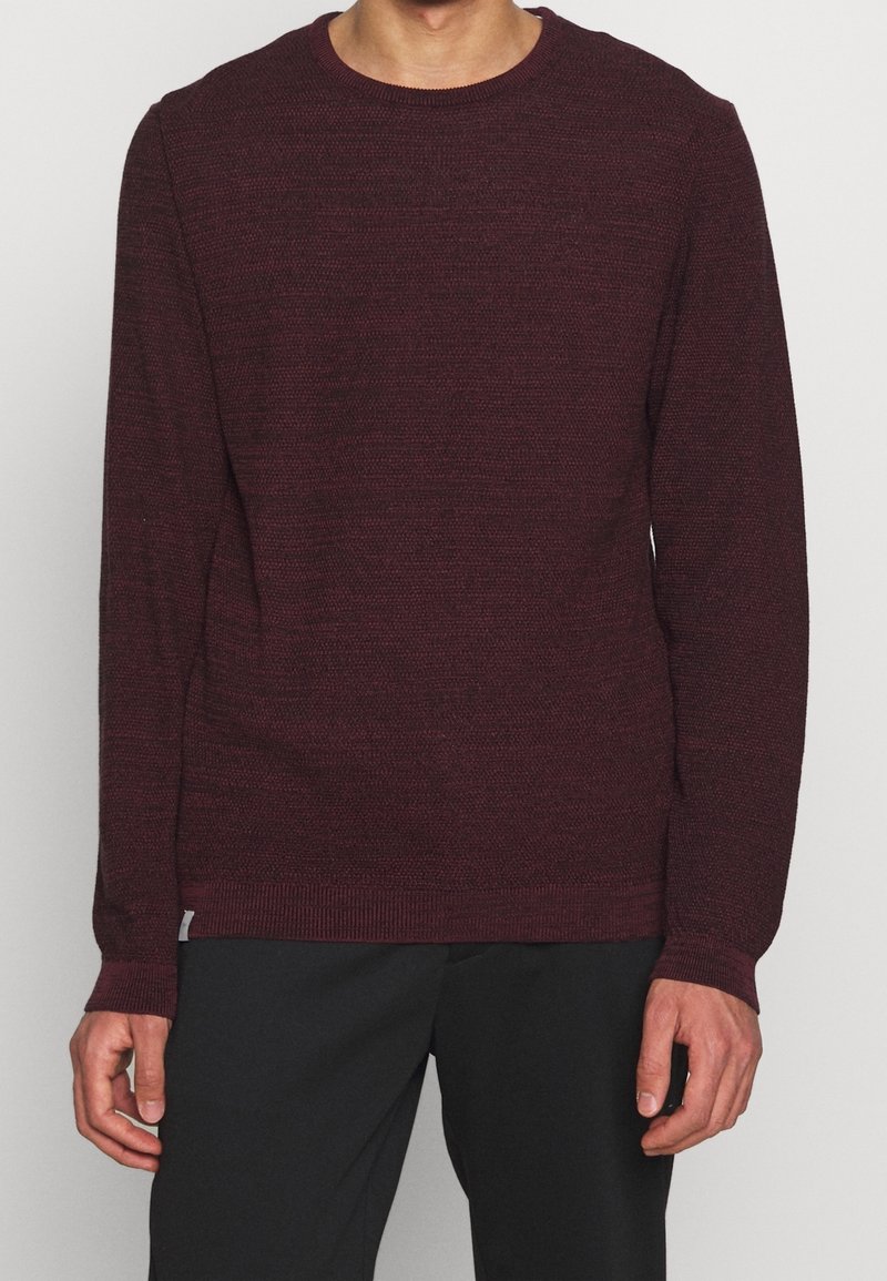 Vínový svetr s kulatým výstřihem v texturovaném pletení, s dlouhými rukávy a žebrovanými manžetami a lemem, nošený přes černé kalhoty.