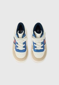 Baskets blanches avec des accents bleus et beige, dotées d'une sangle Velcro et de lacets, avec une tige texturée et un design de bout perforé. Taille marquée comme 24.