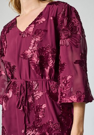 Vinröd klänning med blomsterbroderi, V-ringning, halvgenomskinliga ärmar och knytband i midjan. Har strukturerade, upphöjda blomsterdetaljer.