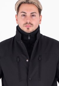 Giacca nera con colletto alto, chiusura frontale con zip e due tasche sul petto. Il tessuto ha una finitura testurizzata e un design aderente.