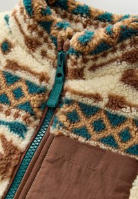 Gros plan sur une veste en polaire montrant une fermeture éclair turquoise, une poche en tissu marron et un motif texturé avec des formes géométriques crème, marron et turquoise.
