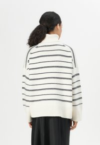 Übergroßer weißer Pullover mit schwarzen Streifen, hoch gerippter Kragen und abgesenkten Schulternähten. Weiche Textur, längerer hinterer Saum.