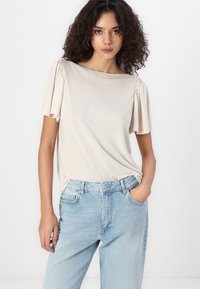 Bežinis trumpomis rankovėmis viršus su plisuotomis rankovėmis, derinamas su šviesiai mėlynomis aukšto liemeniu džinsais. Audinys atrodo minkštas ir šiek tiek tekstūruotas.