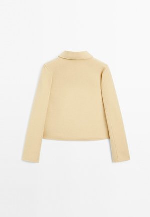 Veste cropped en mélange de laine beige avec des manches longues et un col structuré. Texture lisse, design minimaliste sans fermetures visibles.