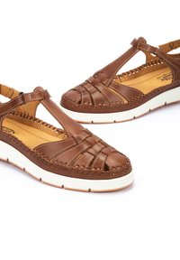 Sandalias de cuero marrón con diseño trenzado, tira en T y suela blanca. Presentan detalles de costura y una plantilla acolchada para mayor comodidad.
