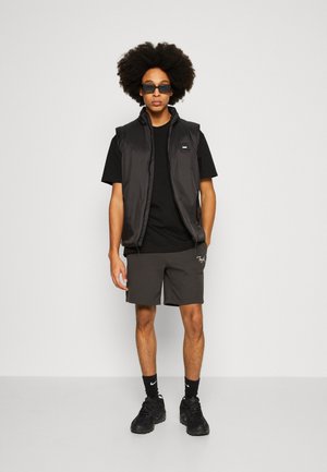 Zwarte mouwloze vest, gewoon zwart T-shirt, grijze shorts met logo en zwarte sneakers. Het model draagt een zonnebril en heeft krullend haar.