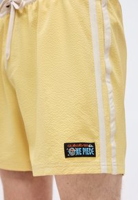 Κίτρινα κοντά παντελόνια με υφή, λευκές πλευρικές ρίγες και μαύρο έμπλαστρο κοντά στο τελείωμα με τα λογότυπα "Quiksilver" και "One Piece".