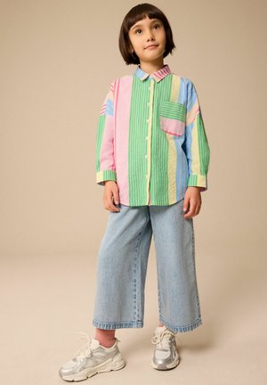 Chemise à manches longues rayée colorée présentant des motifs verts, roses, bleus et jaunes, avec une poche à rabat. Assortie à un jean ample bleu clair et des baskets.
