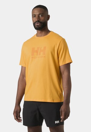 Hombre con barba vestido con una camiseta amarilla de Helly Hansen y pantalones cortos negros con logo HH sobre un fondo gris.