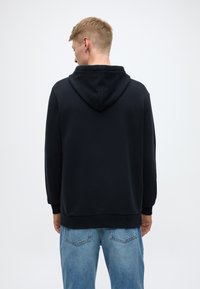 Črna kapucar z dolgimi rokavi, z ohlapnim krojem in rebrastimi manšetami. Nošena s svetlo modrimi jeans hlačami, teksturirana tkanina.