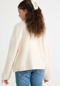 Pige med langt krøllet hår, bundet med en hvid scrunchie, iført en cremet sweater og blå jeans, som står sidevendt mod en ensfarvet baggrund.