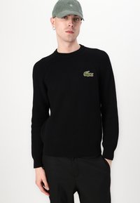 Pull noir en tricot côtelé avec col rond et manches longues, arborant un petit logo crocodile Lacoste en vert et rouge sur la poitrine gauche.
