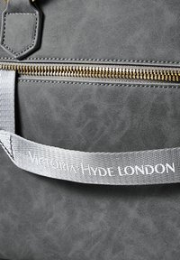 Šedá texturovaná taška se zlatým zipem a bílým látkovým popruhem s nápisem "Victoria Hyde London" v detailním záběru.