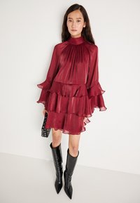 Burgunderfarget ruffled kjole med lange, utsvingte ermer og høy hals. Stoffet har en gjennomsiktig, teksturert finish. Brukt sammen med høye, spisse svarte støvler.